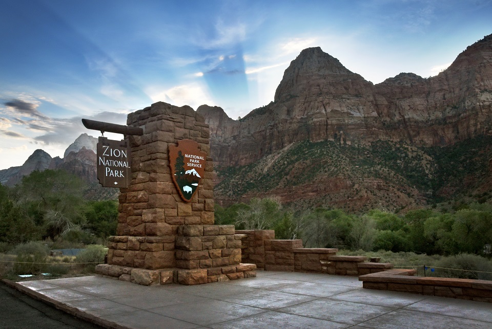 The sun rises behind canyon walls and the Zion National Park entrance sign. 