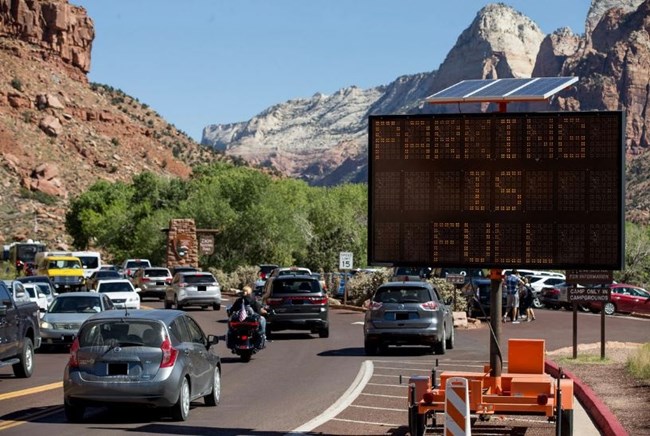 A line of cars wait on the highway outside the park by a sign that says "parking full"