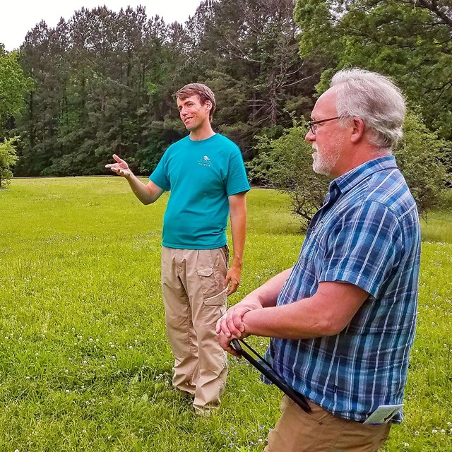 Two gentlemen looking out at a field surrounded by trees with one person speaking