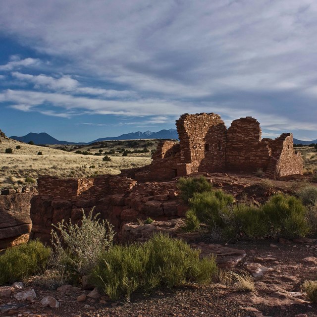 remains of a pueblo surrounded by a desert mountain landscape and cloudy sky