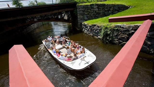 An open boat makes its way down a canal way with people sitting on either side.