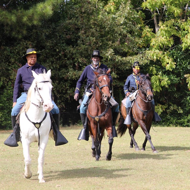 Four men dressed in USCT uniforms ride horses 