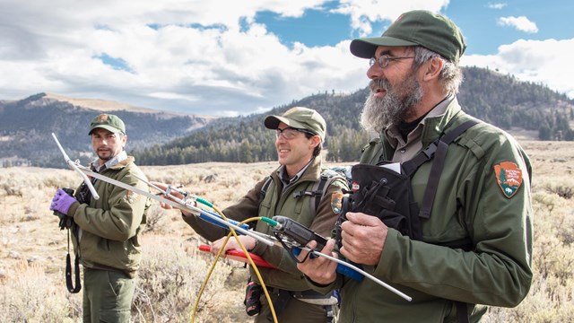 Three uniformed park employees testing telemetry gear in a meadow.