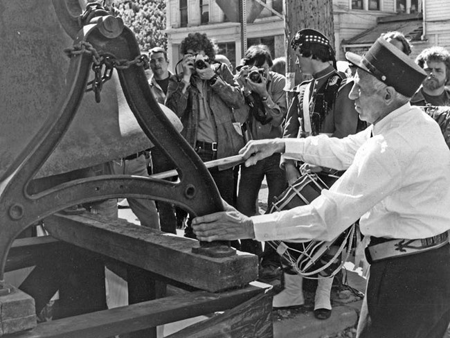 black and white image of man standing near a large metal object, surrounded by a crowd of people